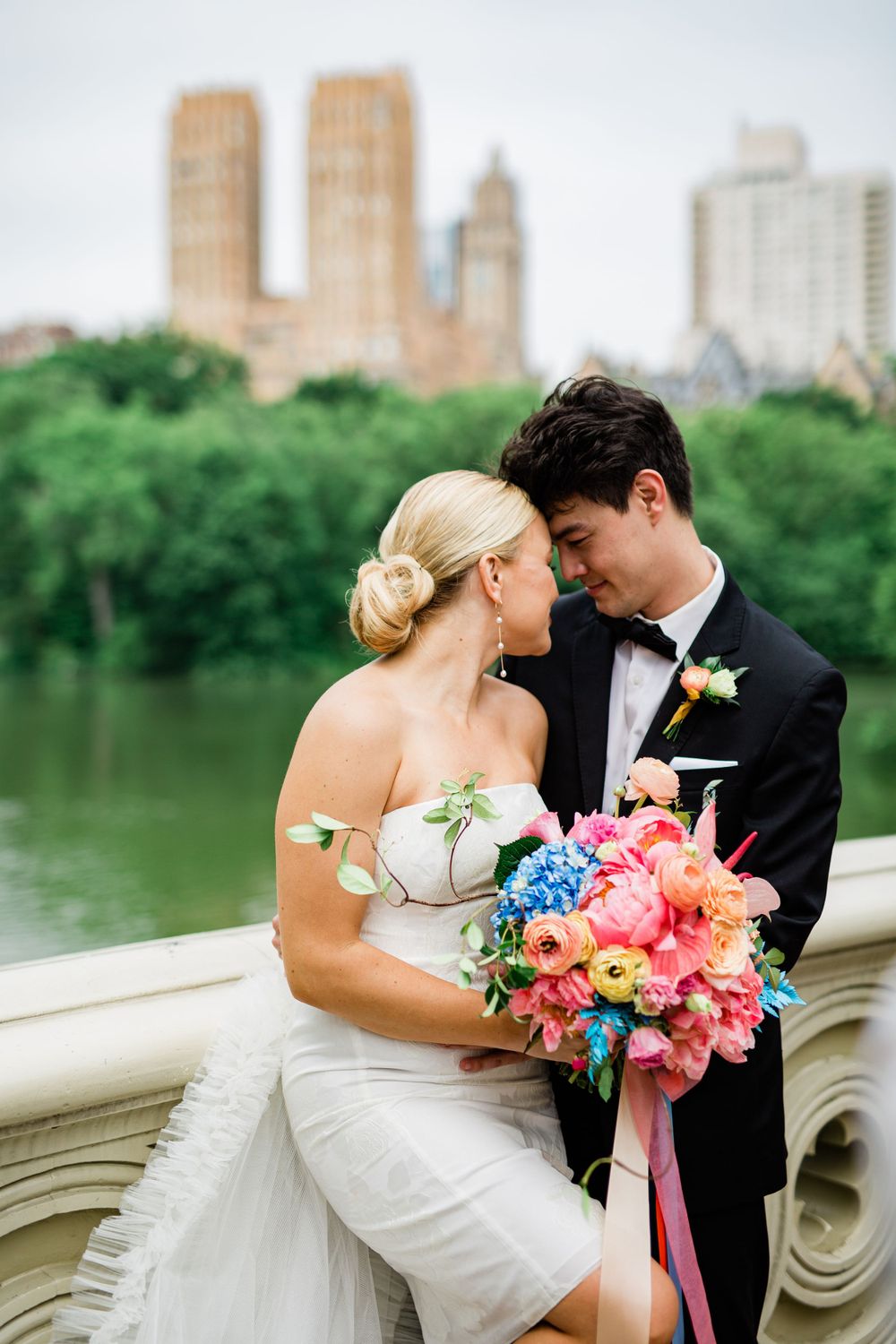 A tender wedding moment shared on a stone bridge with a colorful bouquet and city backdrop.