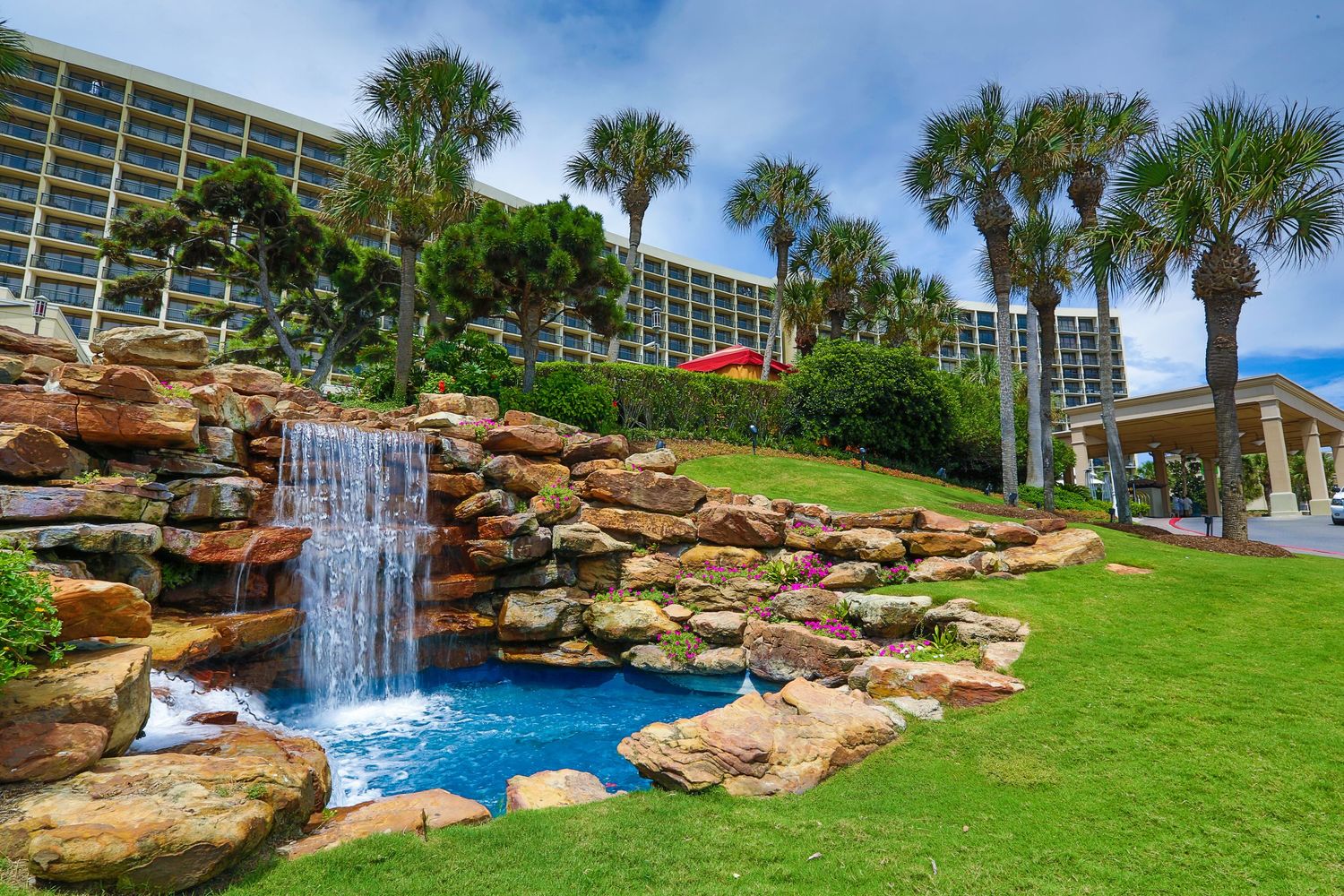Luxurious resort waterfall feature cascading over natural rock formations with palm trees and buildings in background.