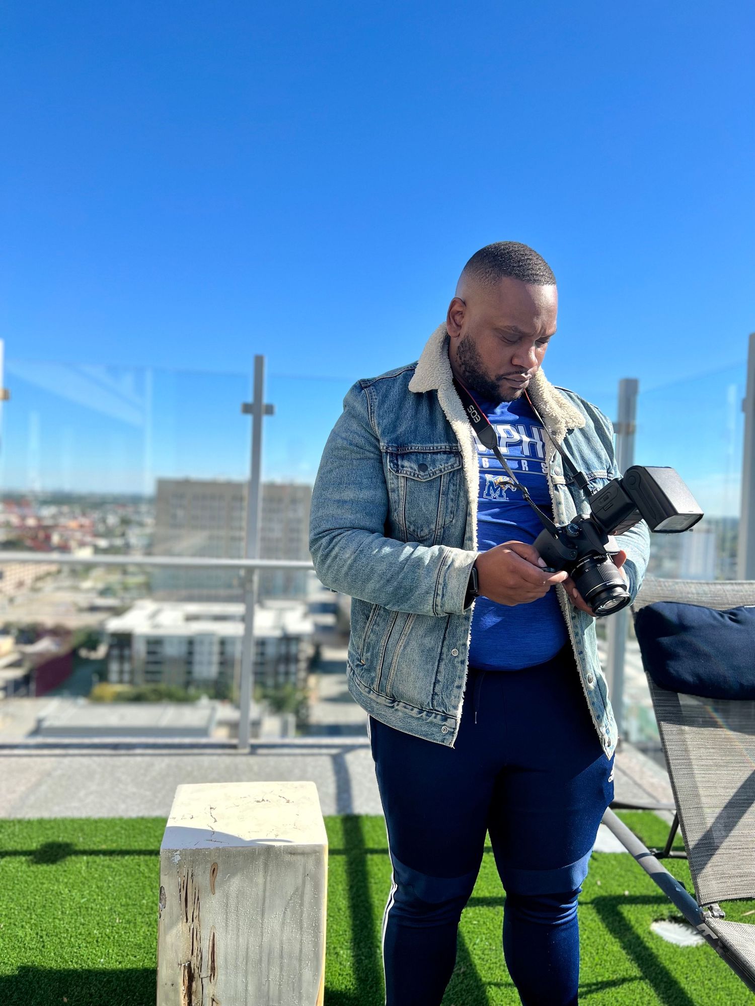 A photographer checks camera equipment on a rooftop with artificial turf and city views in background.