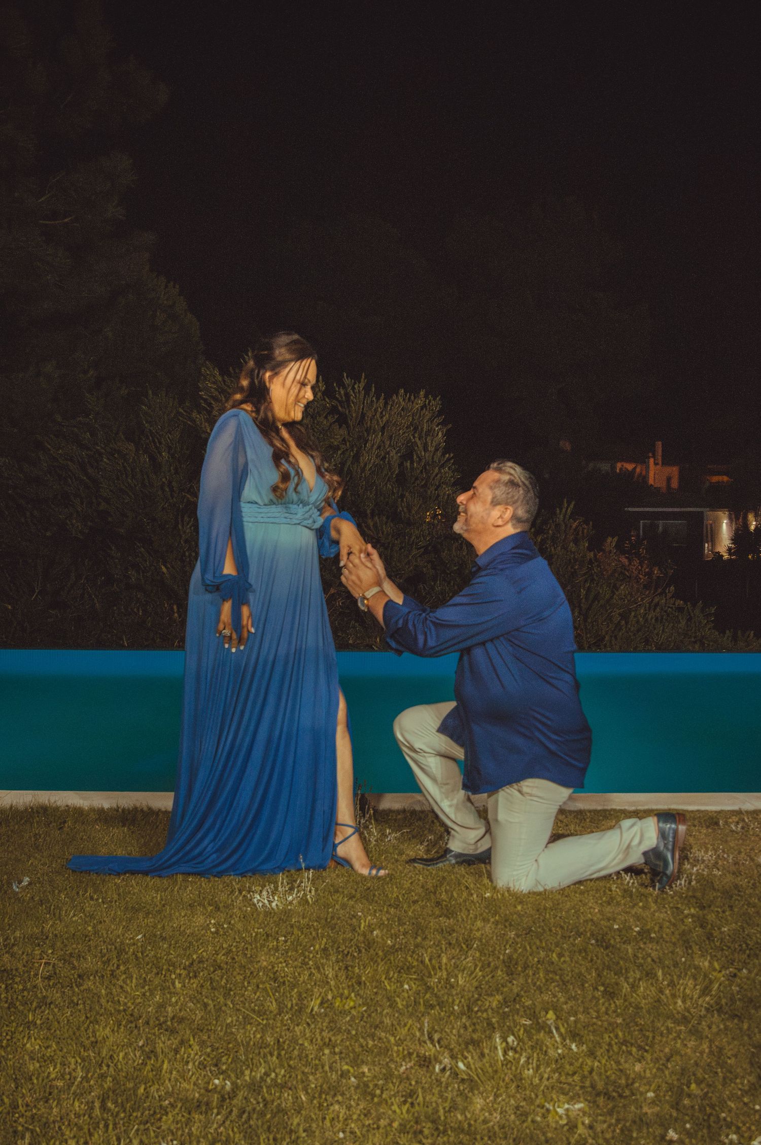 A nighttime proposal scene on grass near a blue-lit swimming pool.