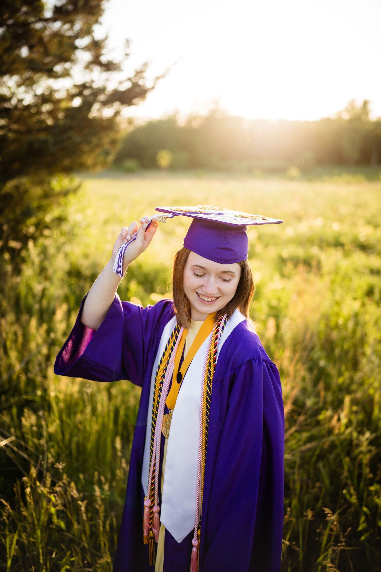Graduate in purple cap and gown poses in golden sunset field.