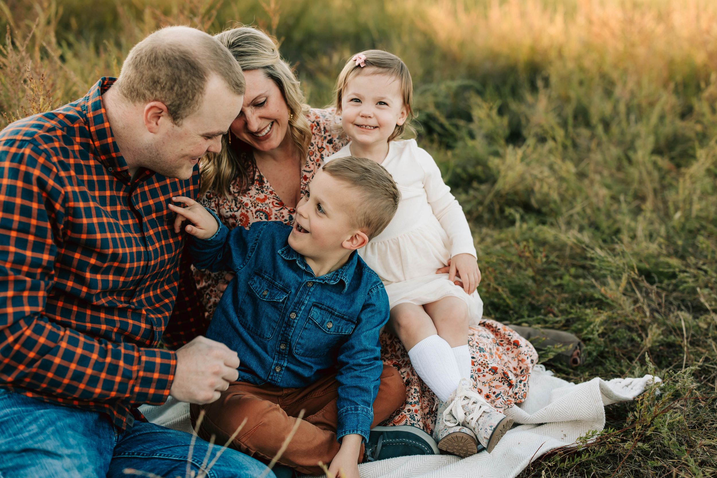 Family shares candid moment together while sitting in golden field at sunset.