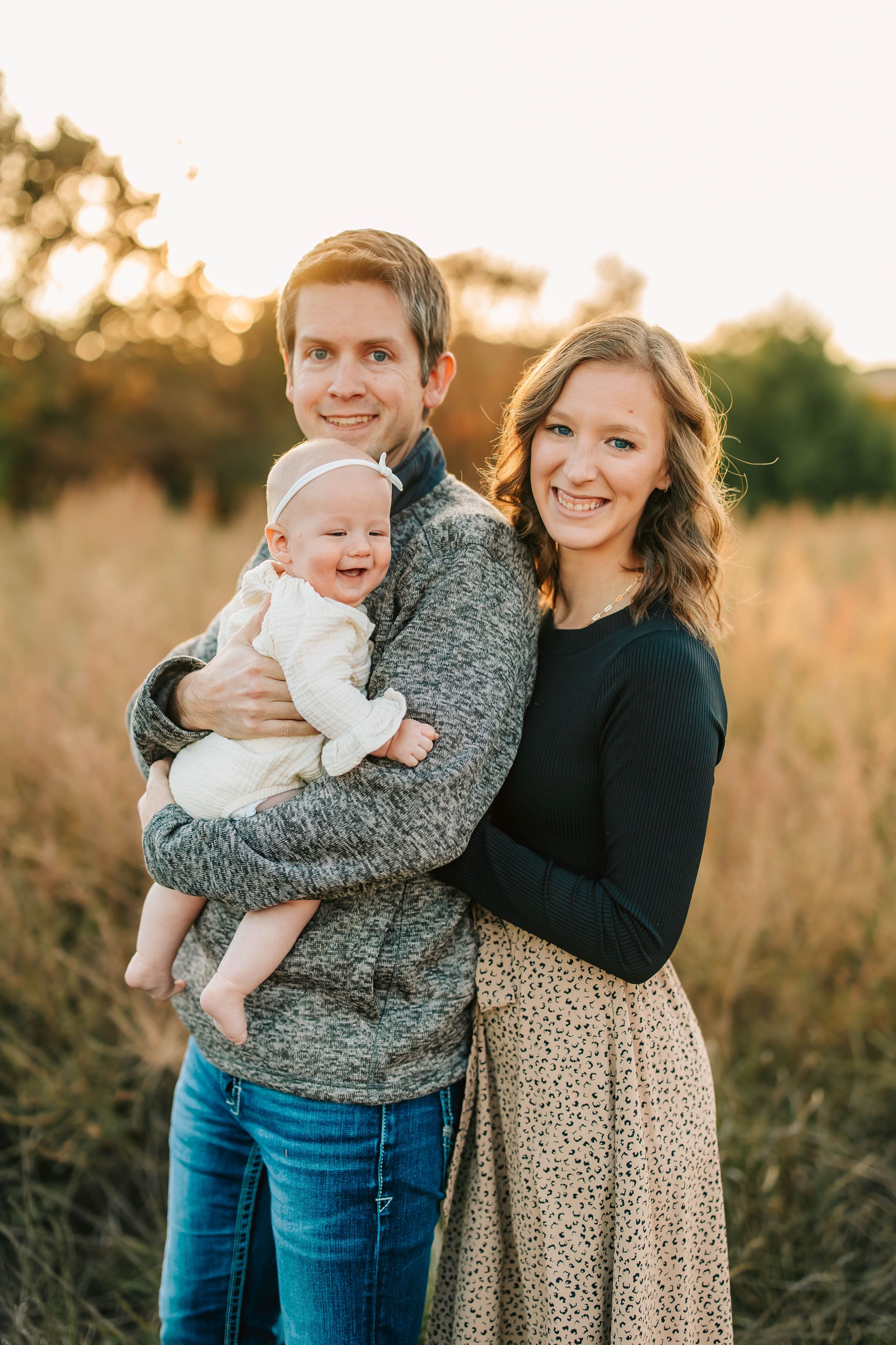 Happy couple poses with their baby in golden field at sunset for heartwarming family portrait.