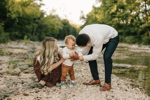 Parents kneeling with toddler on rocky riverbank during outdoor family photoshoot.