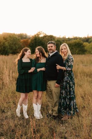 A group poses together in a field at sunset wearing coordinated outfits with green dresses and white boots.