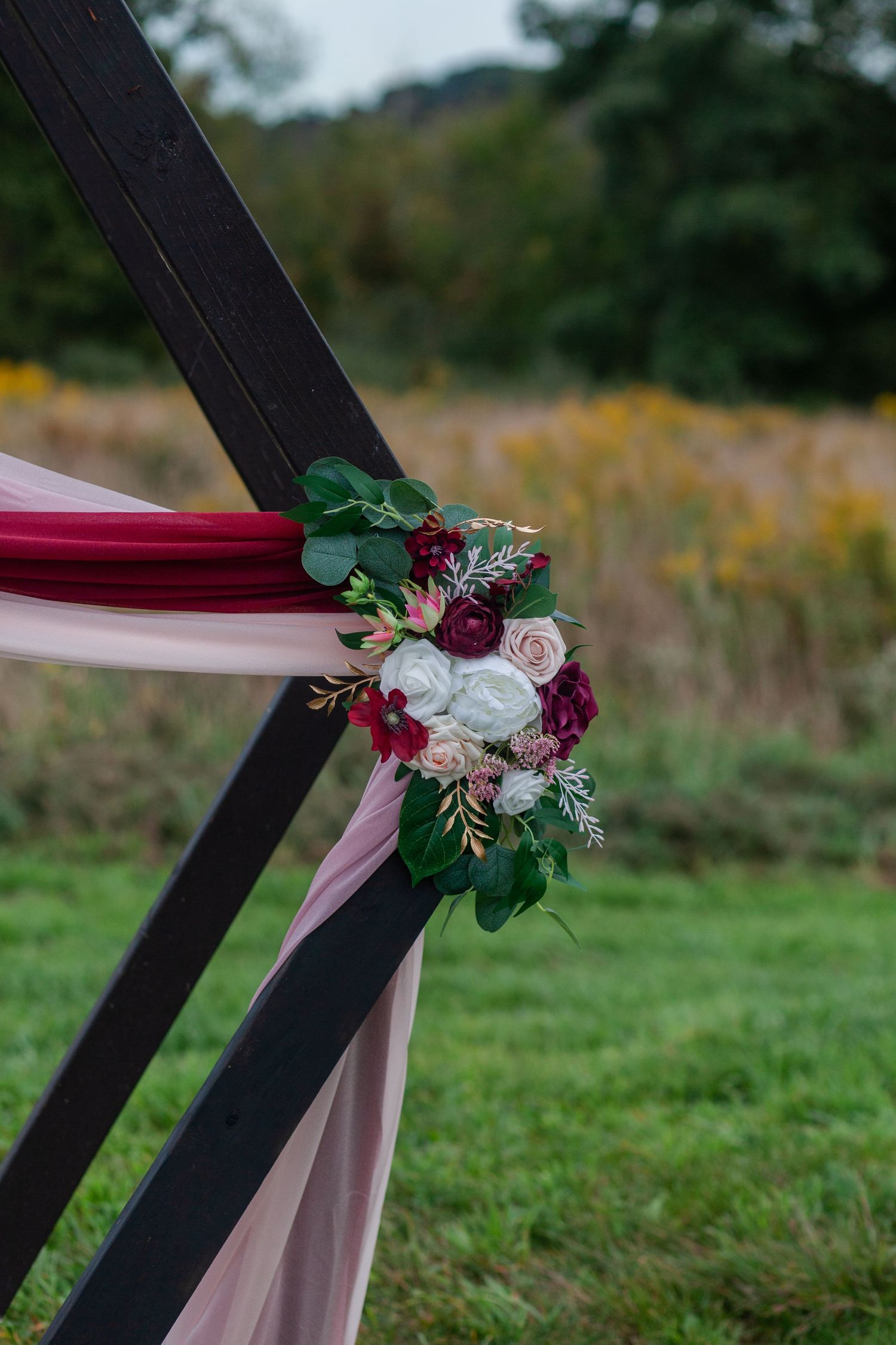 Elegant floral arrangement with burgundy and white flowers adorns a black wedding arch draped with pink fabric.
