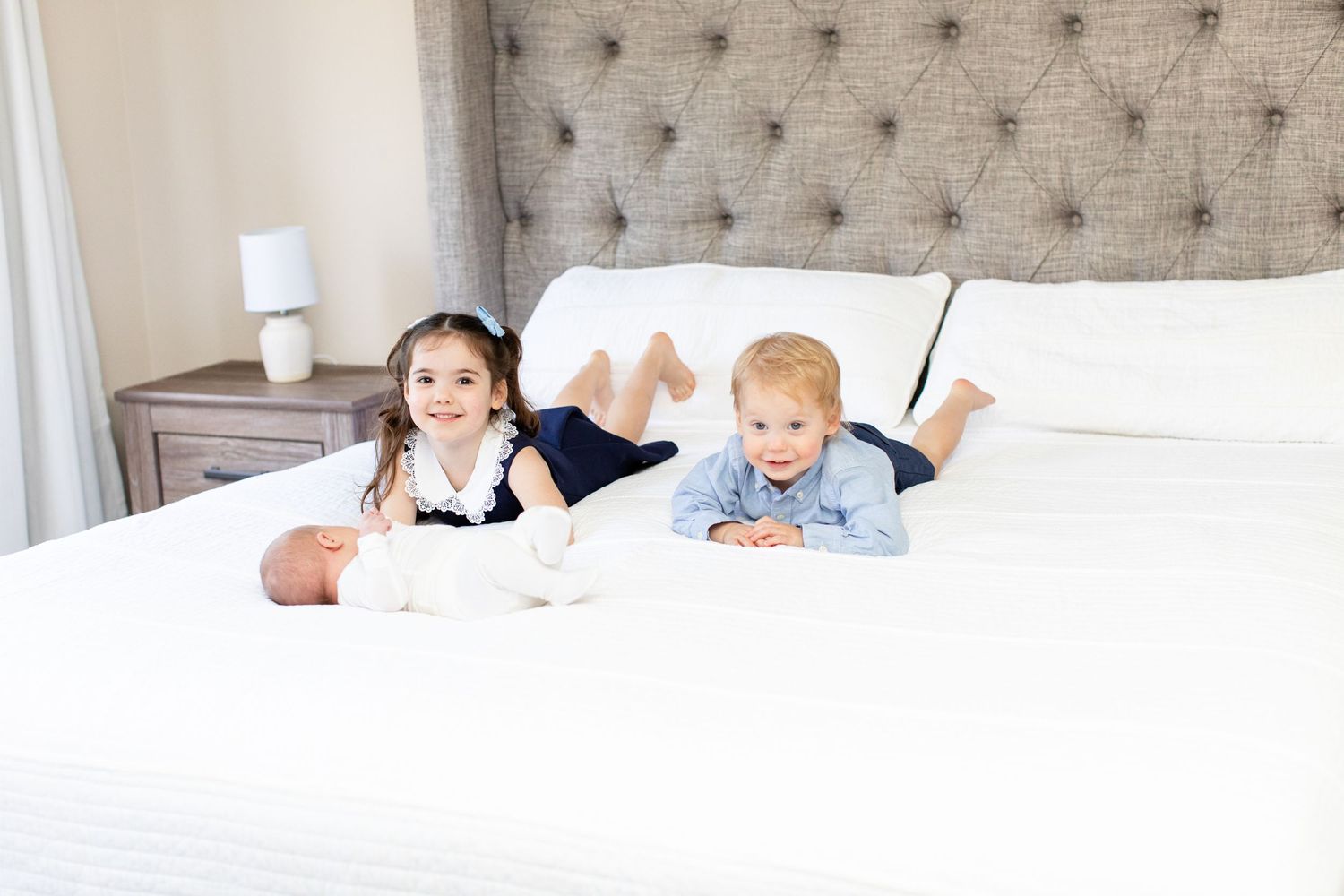 Two young children relaxing on a white bed with tufted gray headboard.