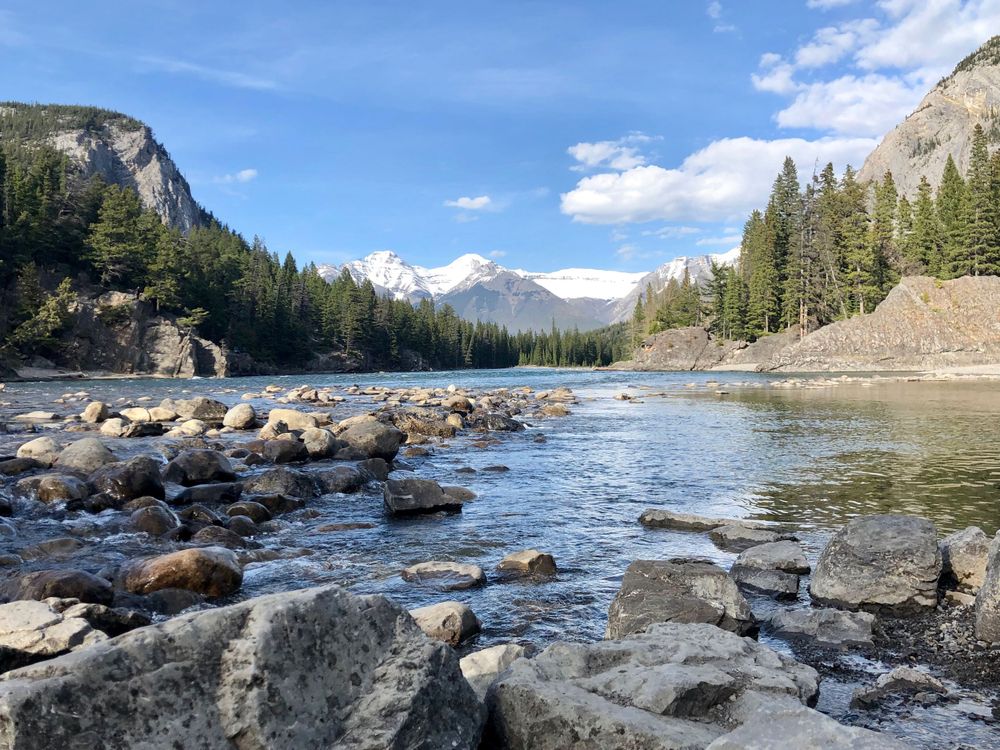 Beautiful mountain landscape with rivers and rocks surrounded by evergreen forest and snow-capped peaks.