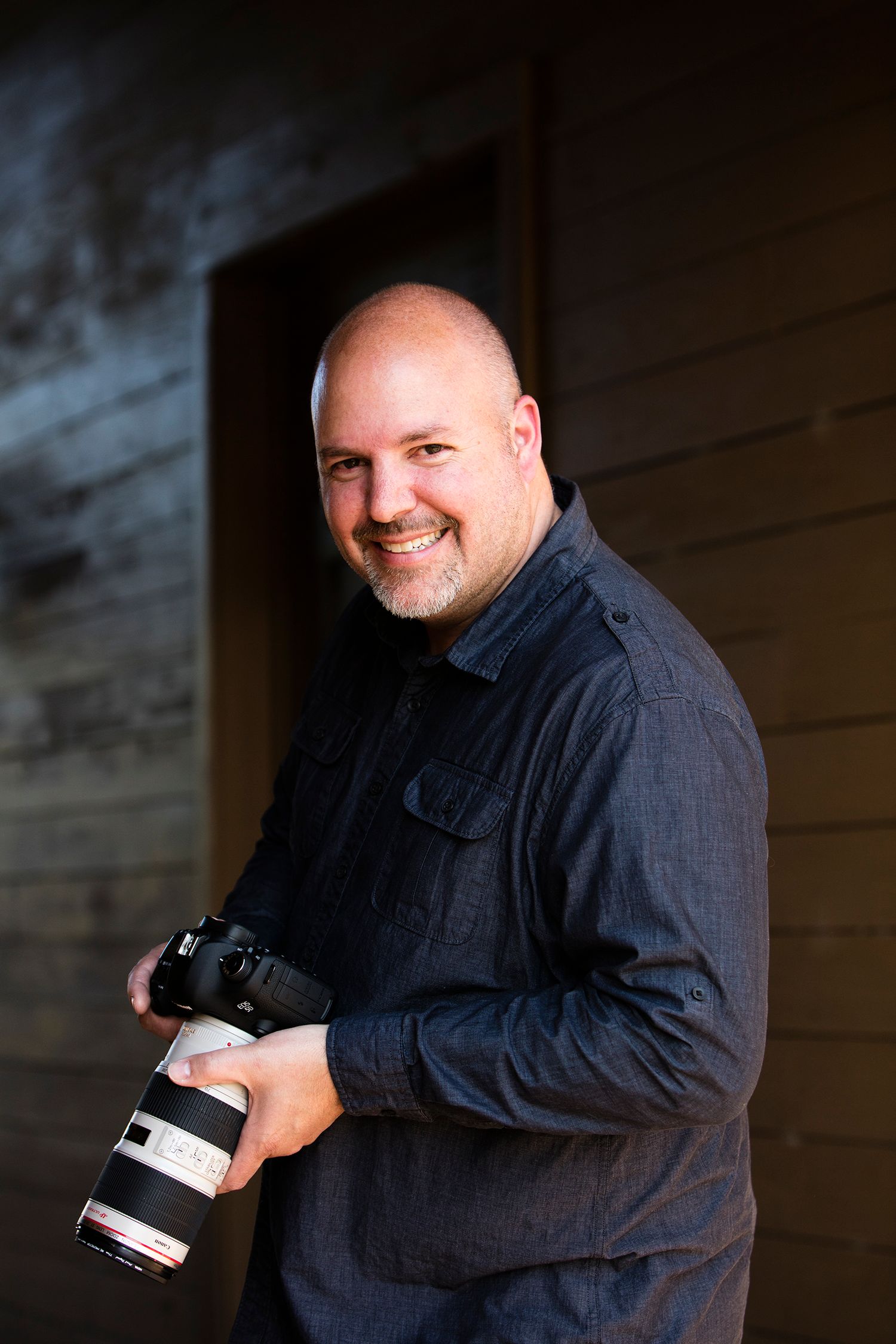 Photographer in a navy shirt holds a professional camera with telephoto lens against a dark background.