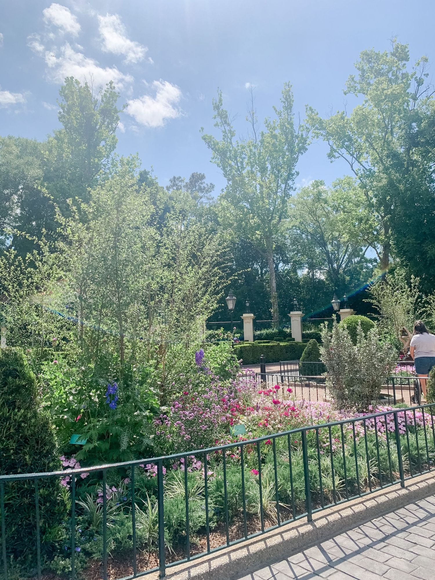 Garden walkway with black metal railings surrounded by lush trees and flowering plants.