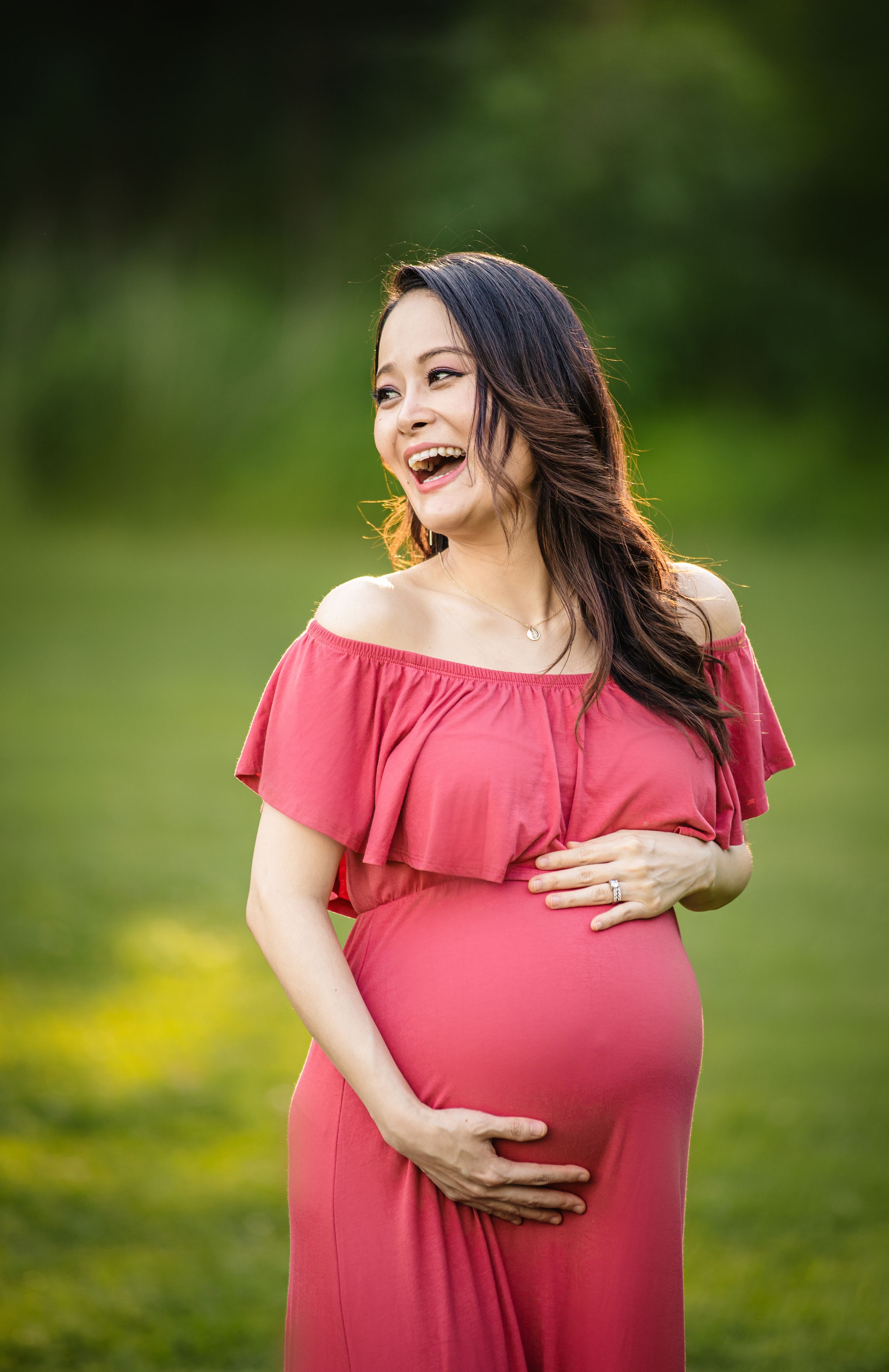 Expectant mother in flowing red off-shoulder dress laughs joyfully in outdoor garden setting.