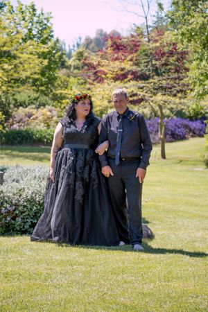 A couple in formal attire poses on a lawn with colorful autumn foliage in the background.