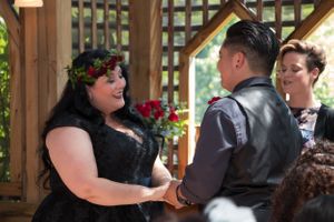 A wedding ceremony takes place under a wooden gazebo with red flowers in the background.