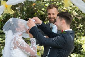 Groomsmen help lift and arrange the bride's veil during an outdoor wedding ceremony.