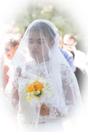 A bride in lace holds yellow flowers under her veil during an outdoor ceremony.