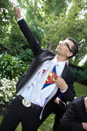 Groomsman reveals Superman costume under suit jacket in playful garden pose.