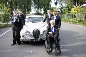 Wedding party members pose with a classic white Jaguar car on a tree-lined road.