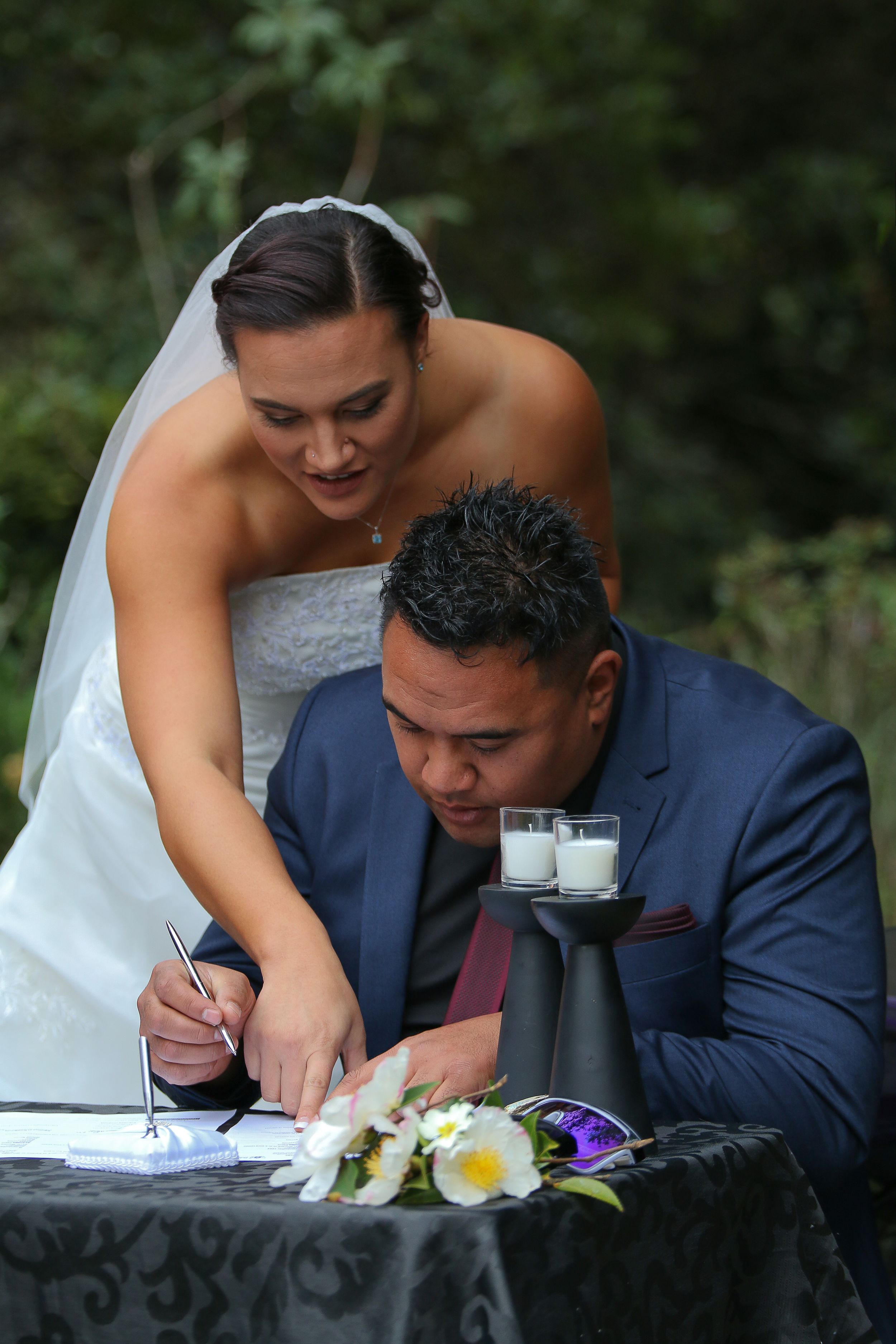 A couple signs their marriage license at a black-clothed table with floral decorations.