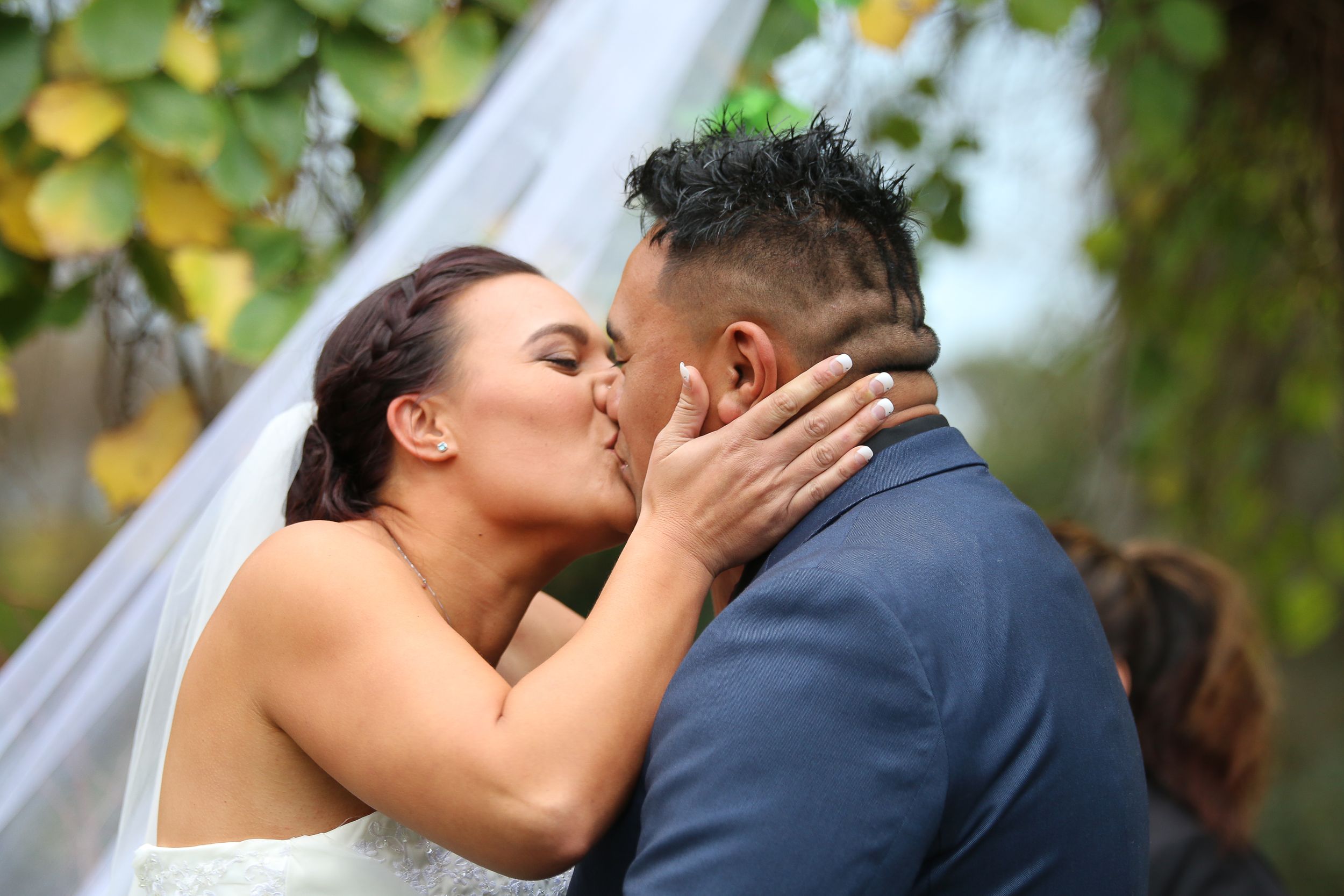 A romantic wedding kiss moment captured outdoors with natural greenery in the background.
