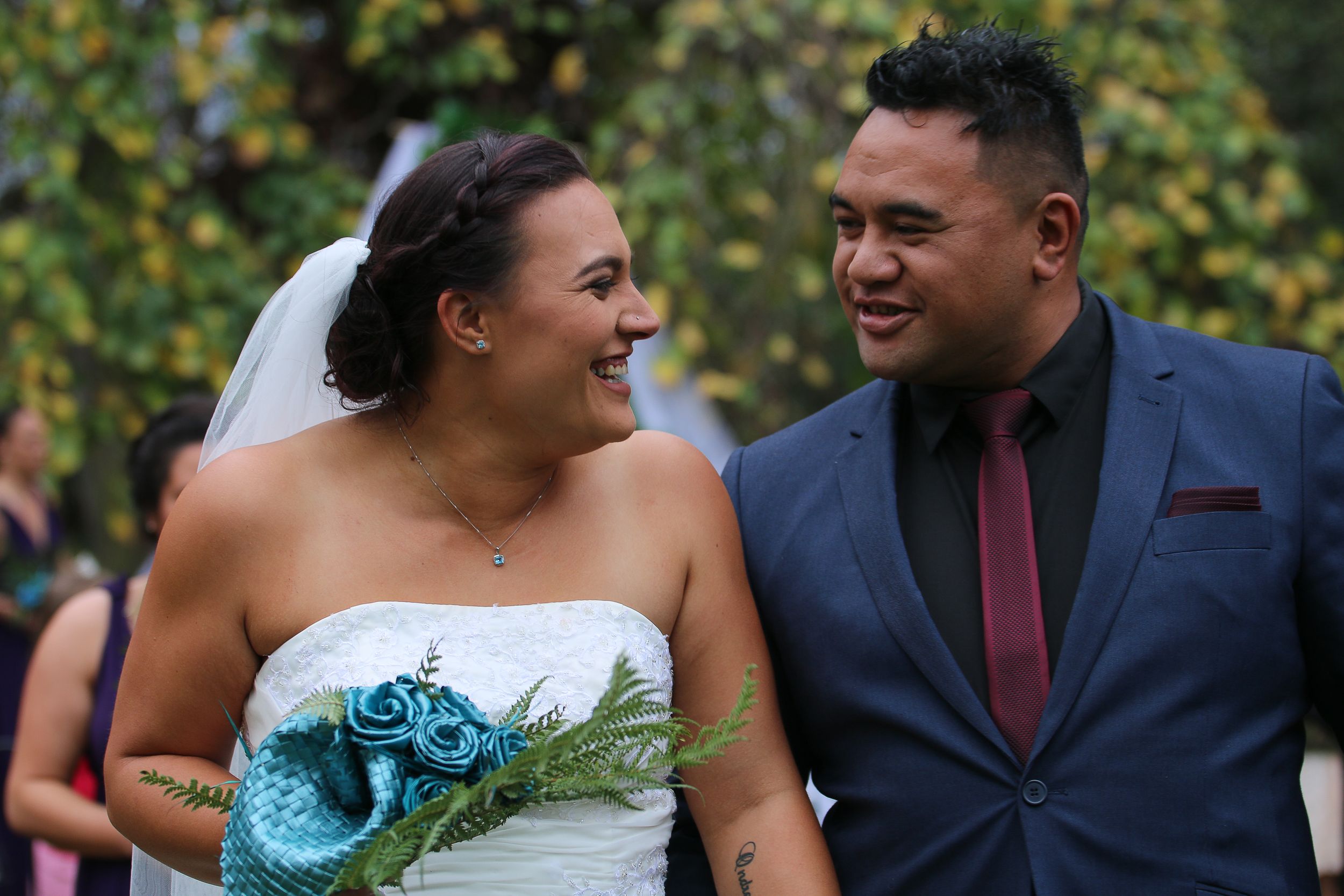 Newlyweds share laughter during outdoor wedding ceremony with bride holding turquoise bouquet.