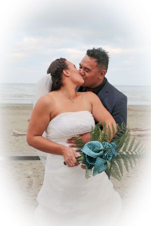 Romantic beachside wedding portrait with couple kissing as waves crash behind them.