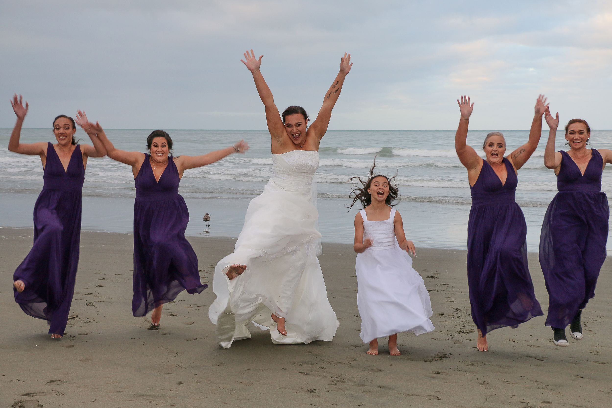 Bridal party jumping joyfully on beach with bridesmaids in purple dresses.