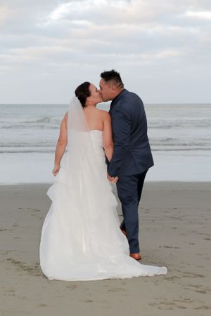 Newlyweds share intimate kiss on beach with flowing white wedding dress and ocean backdrop.