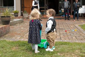 Two small children in formal wear playing on grass near brick pavement.
