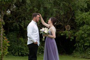 A couple shares a playful moment outdoors as a bridesmaid in purple adjusts something on the groom's face.