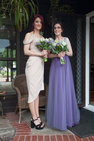 Two bridesmaids in elegant dresses holding floral bouquets pose on a porch.