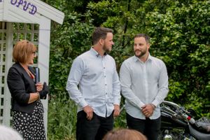 Three people gathered near a white garden arbor during an outdoor event.