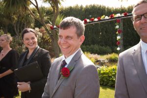 Wedding party members smile outdoors under palm trees with decorative arch.