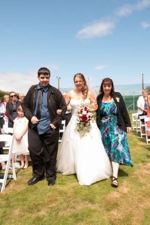 A wedding party stands on a grassy lawn with white folding chairs and blue skies in the background.