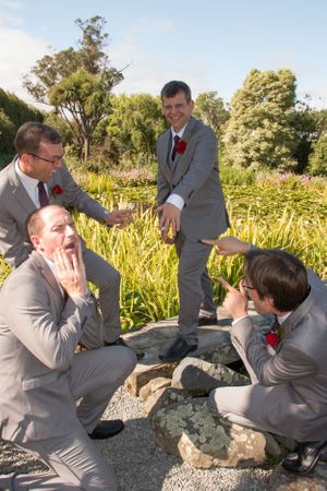 A group in grey suits gathers around rocks in a sunny garden setting during a wedding.