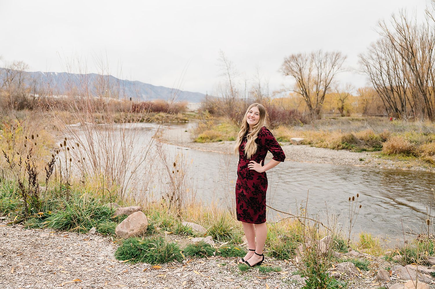Woman in burgundy dress poses by scenic riverside with mountains and autumn foliage in background.