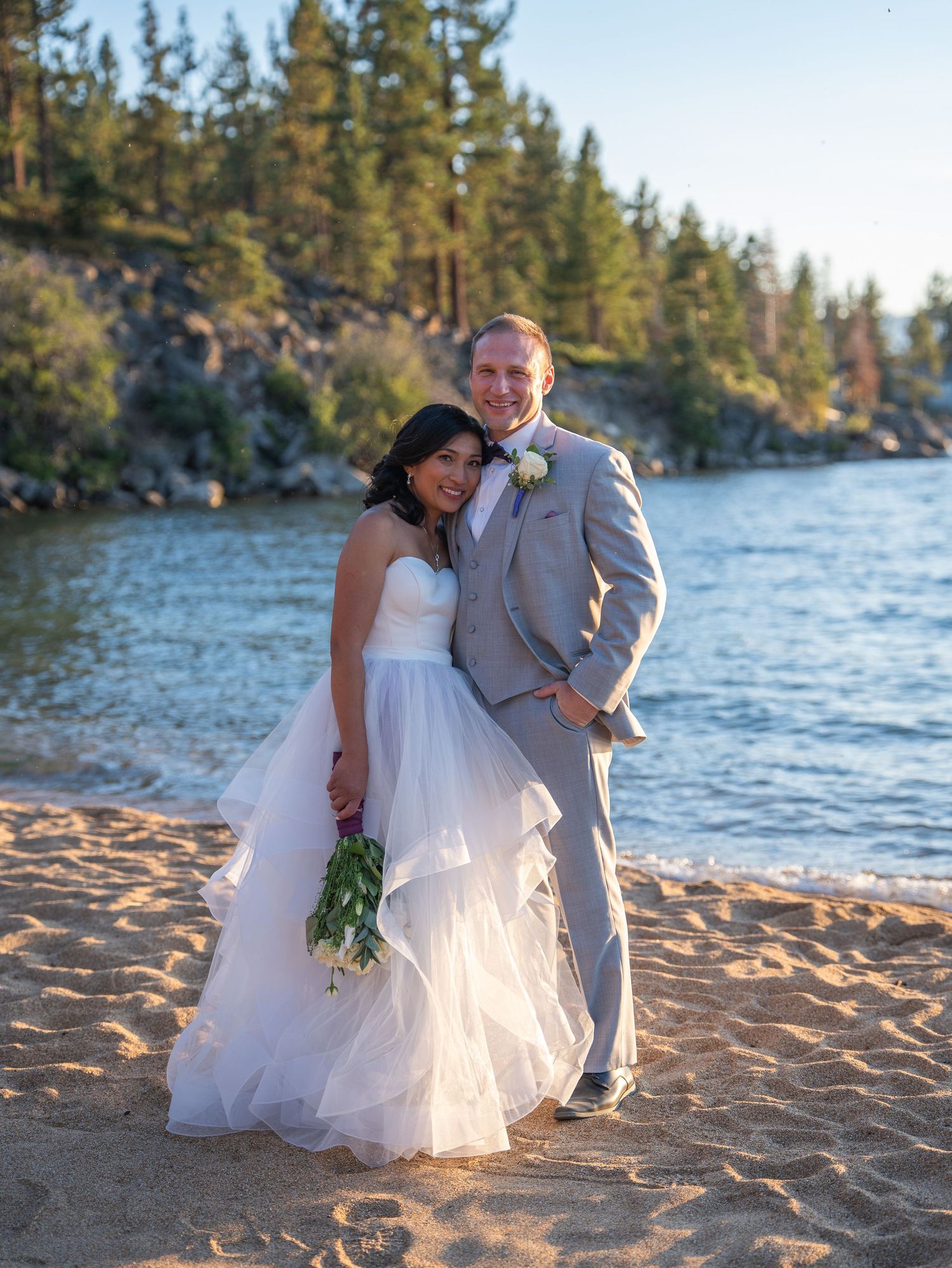 A couple poses on a sandy beach at sunset with pine trees and Lake Tahoe's blue waters in the background.