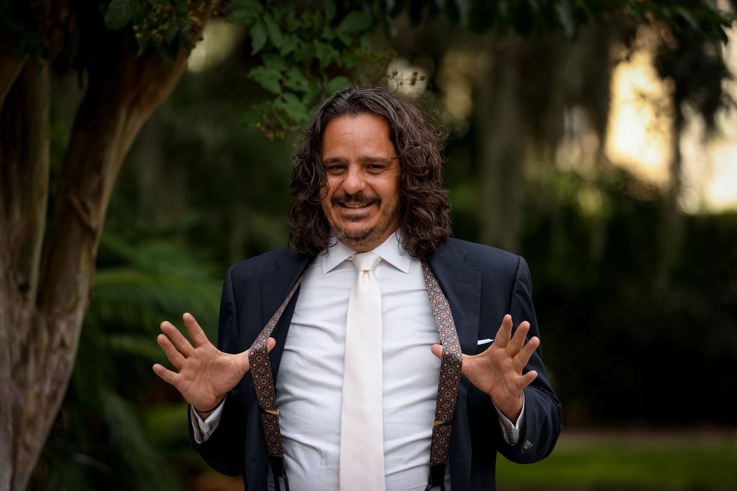 A joyful person wearing a formal suit gestures expressively outdoors.