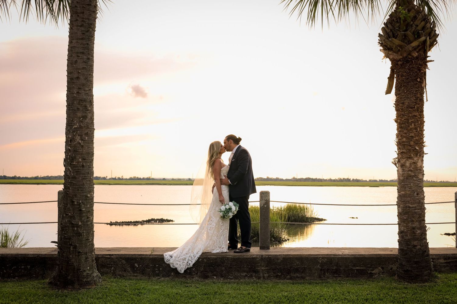 A romantic sunset silhouette of a couple in wedding attire between palm trees by the water.
