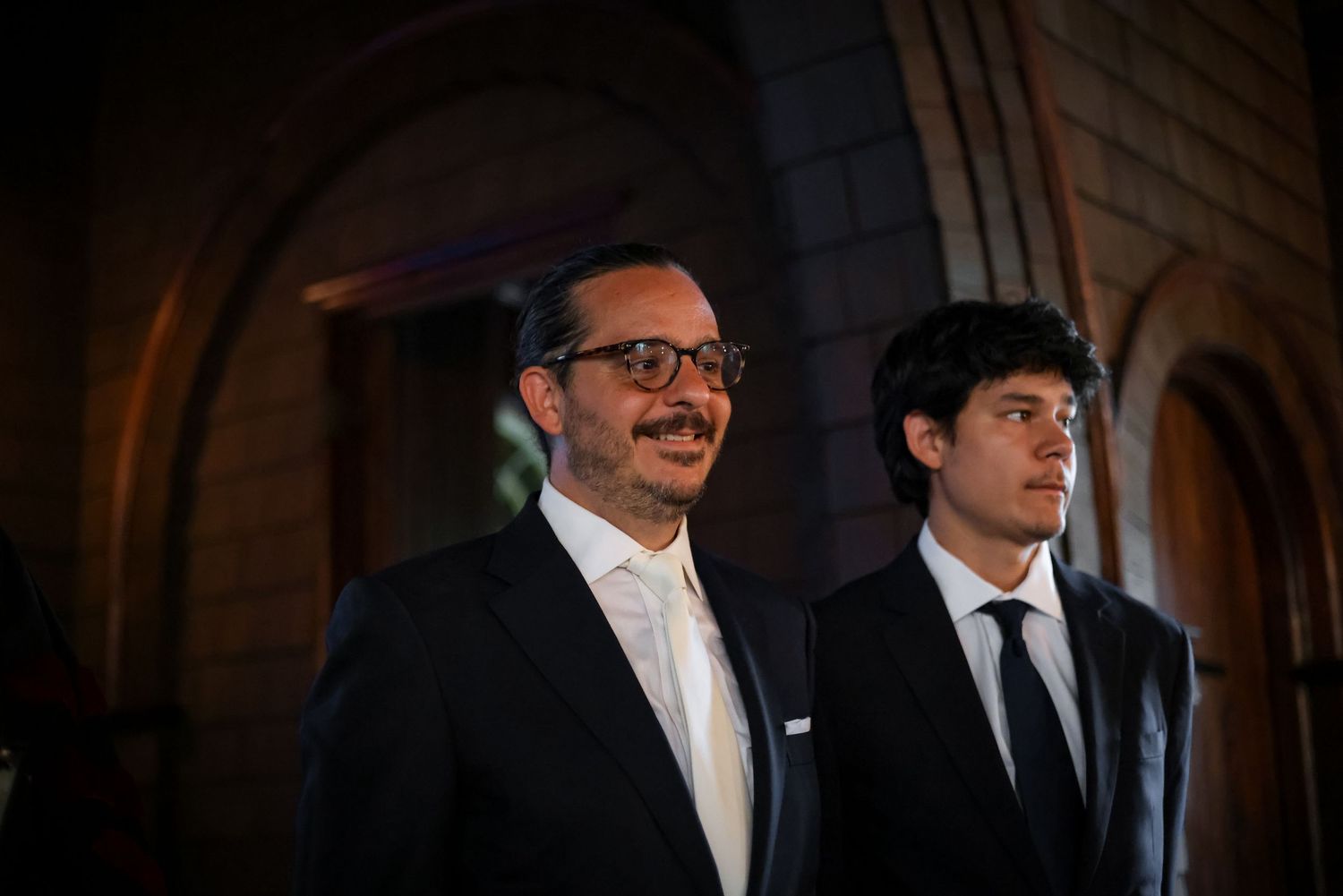 Two people in formal attire stand together in an dimly lit architectural setting.