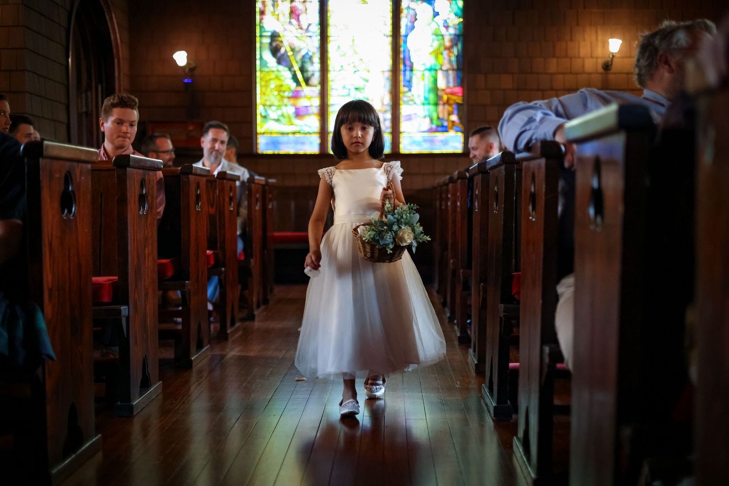 A young attendant in white dress carries flowers down a church aisle with stained glass windows.