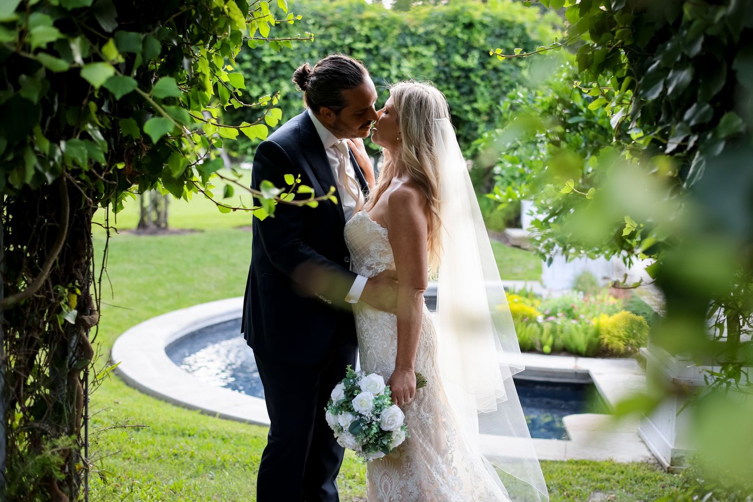 A romantic wedding couple shares a kiss among lush greenery near a garden fountain.