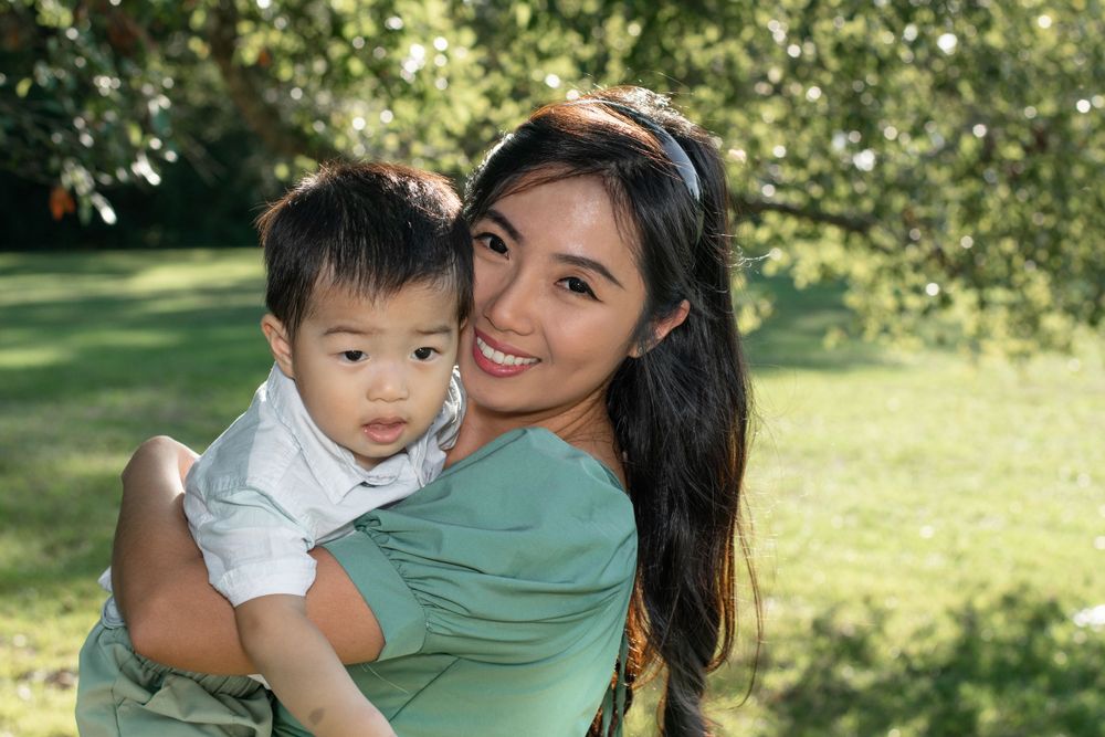 A woman in a green dress holds a small child while smiling in a sunny outdoor setting.