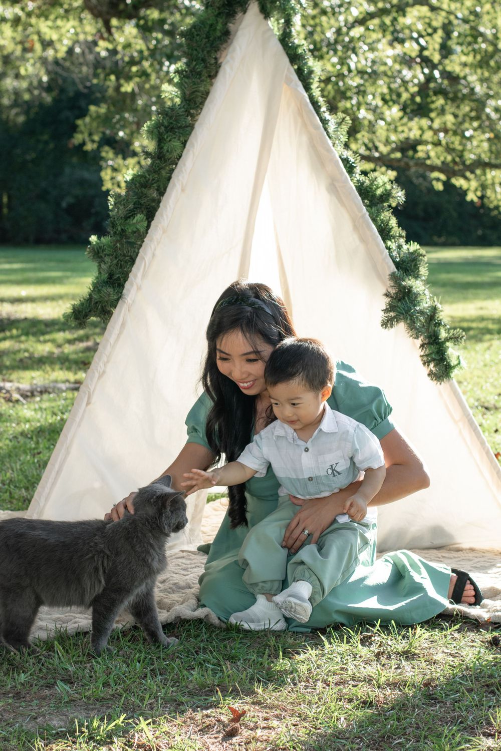A peaceful scene with a grey cat joining a family by a white teepee decorated with greenery in a garden setting.