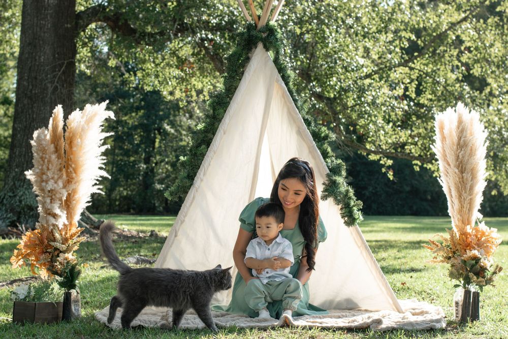 A woman and child sit by a teepee tent with pampas grass decorations and a black cat.