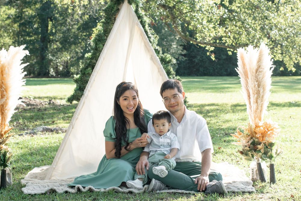 A family sits together on a blanket by a decorated teepee tent in a park setting.