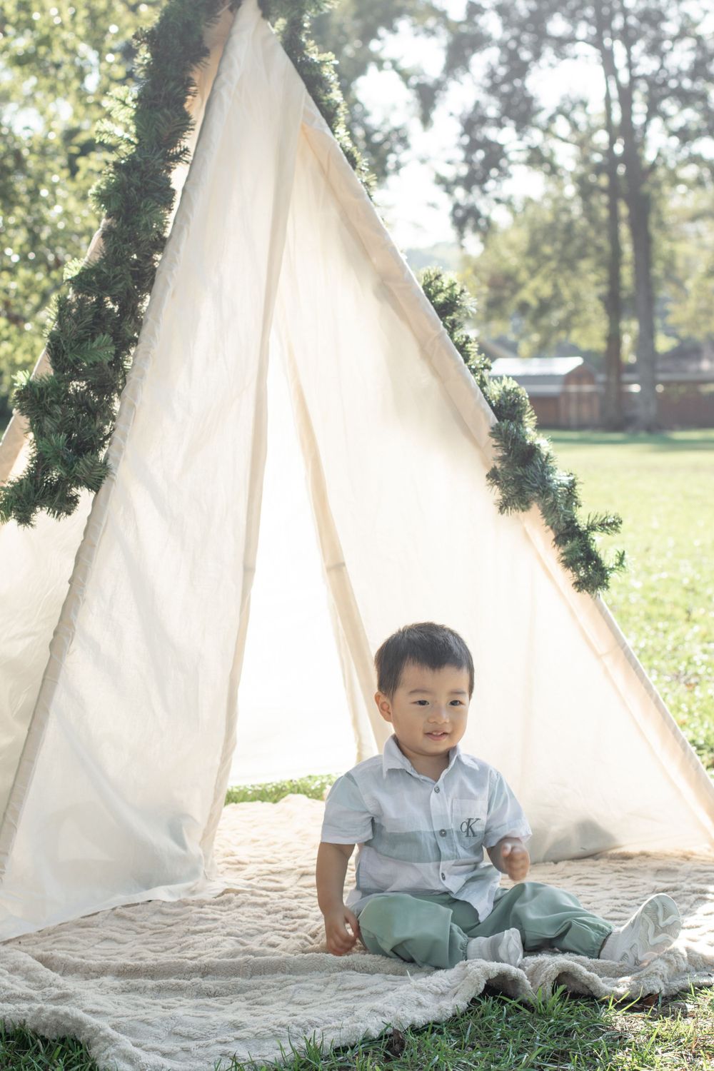 A child sits contentedly on a blanket in front of a white teepee decorated with greenery in an outdoor setting.