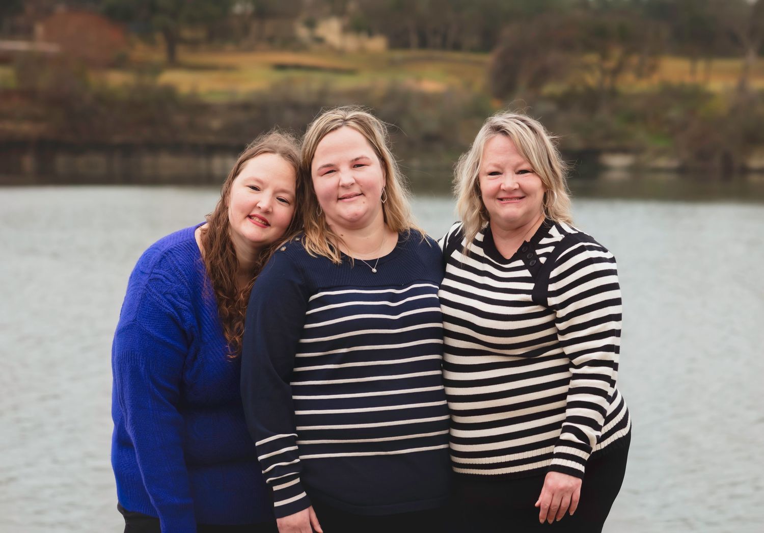 Three women in striped and solid sweaters stand together smiling by a waterfront on an overcast day.