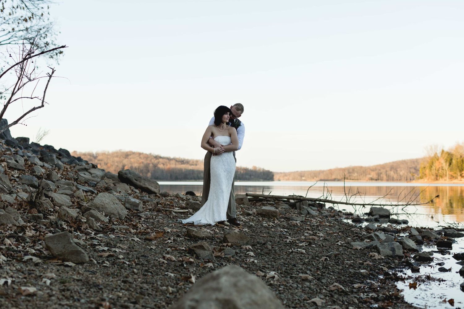 Bride and groom embracing on a rocky lakeside shore at golden hour.