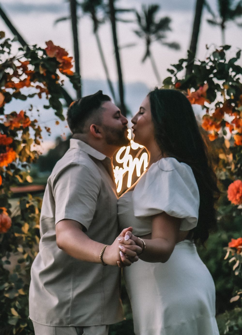 A romantic sunset photoshoot with a couple in white attire surrounded by orange flowers and palm trees.