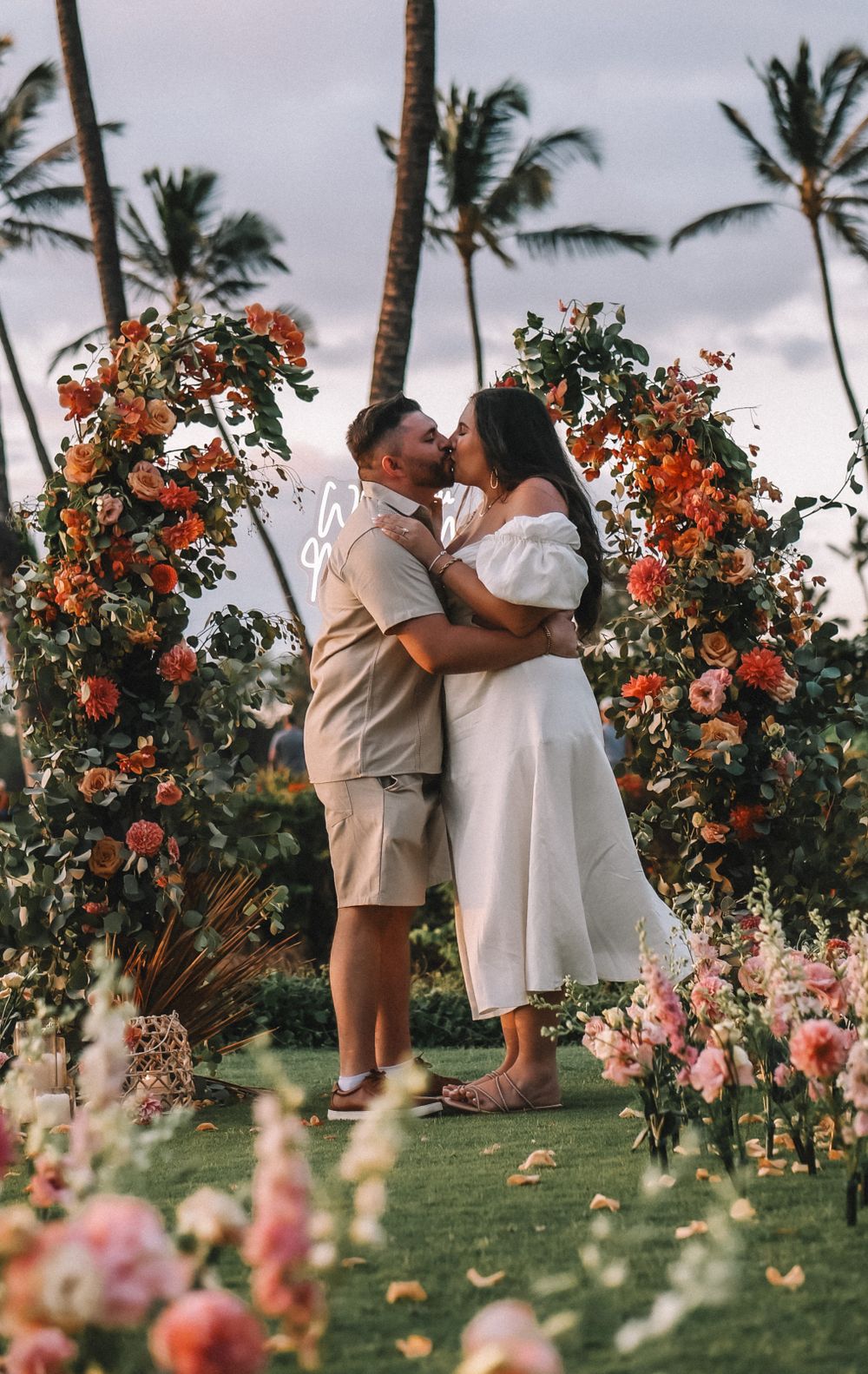 Intimate wedding moment under a coral and pink floral arch on a Hawaiian beach.